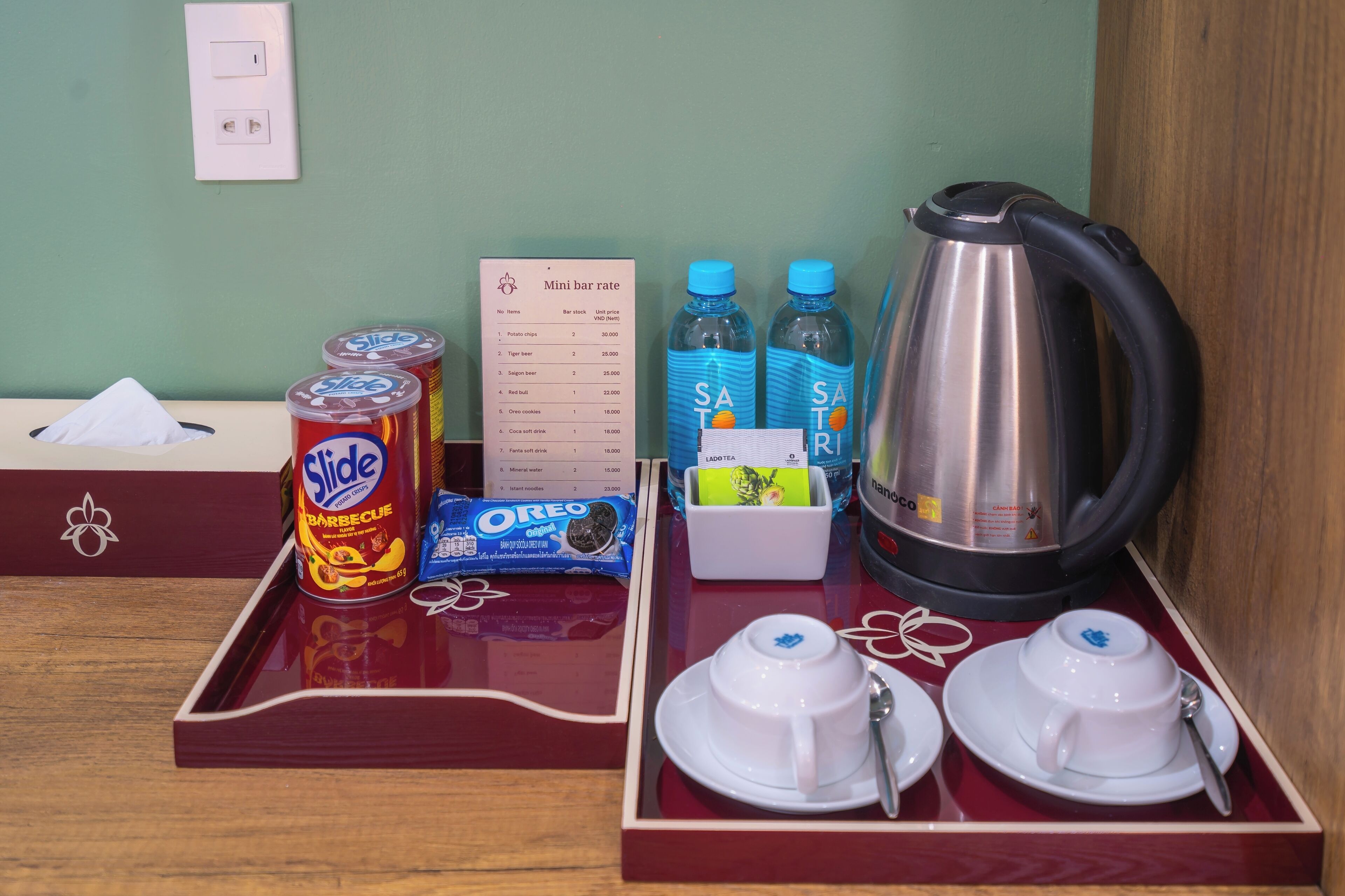 A mini bar setup with a tray containing a can of Slide barbecue sauce, Oreo cookies, a mini bar rate sign, bottled water, and a kettle.