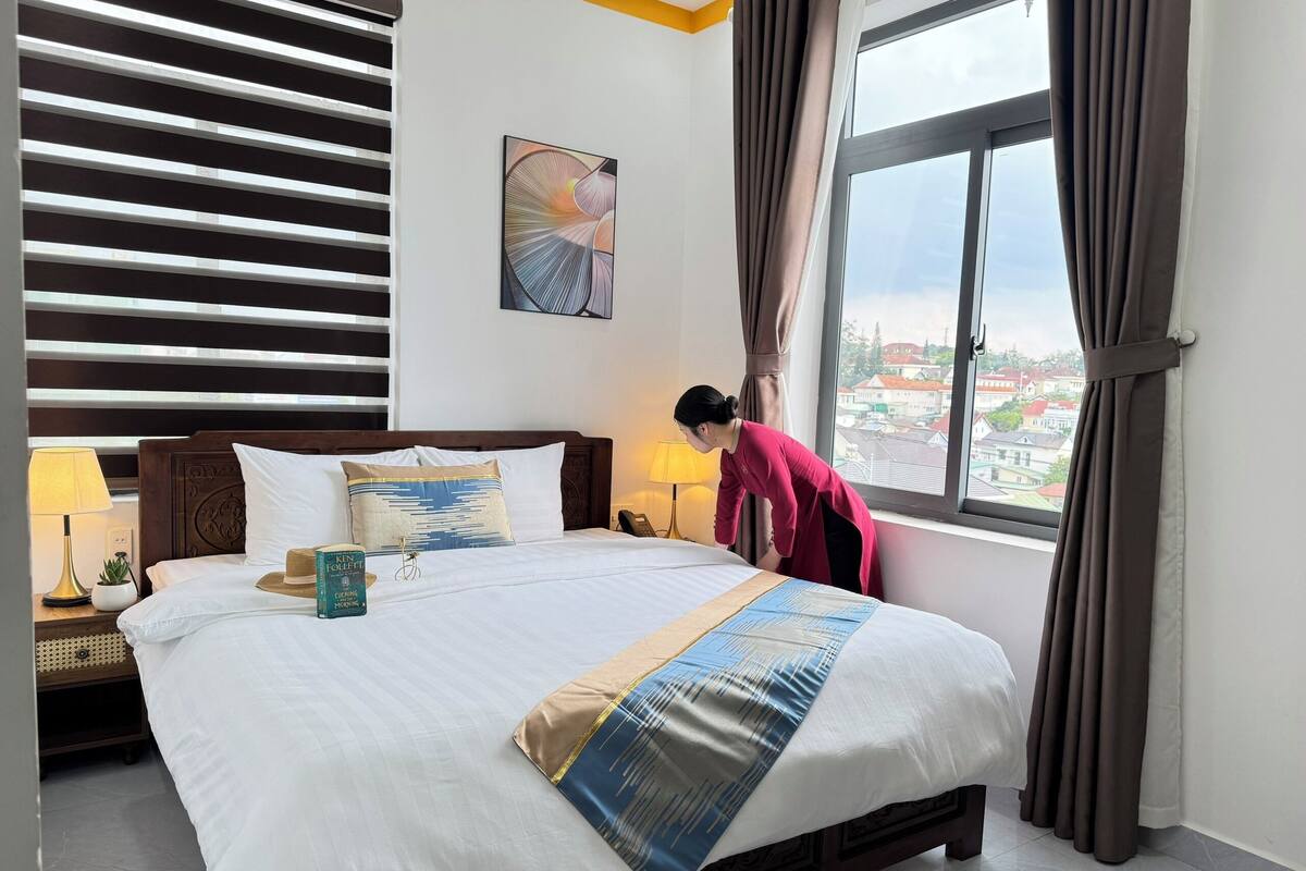 A hotel room with a large bed, a nightstand with a lamp, a window with curtains, and a city view.