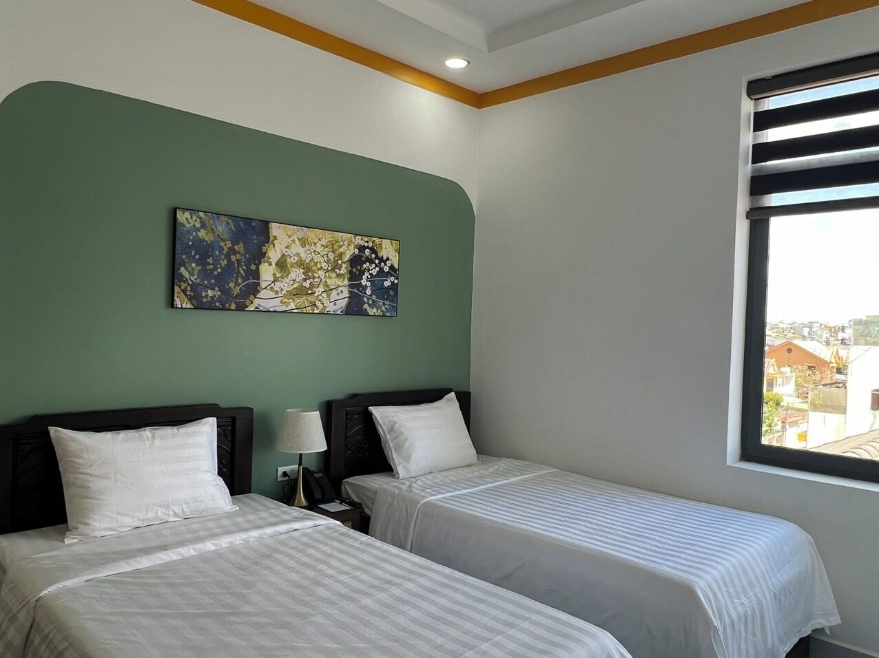 A hotel room with two beds, a nightstand with a lamp, a painting on the wall, and a window with a view of buildings.