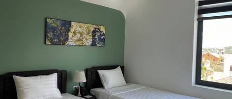 A hotel room with two beds, a nightstand with a lamp, a painting on the wall, and a window with a view of buildings.
