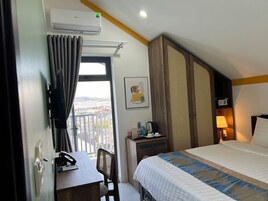 Habitación de hotel con cama, escritorio y silla, ventana con cortinas, aire acondicionado de pared, cuadro enmarcado, mesita de noche con lámpara y vista a la ciudad.