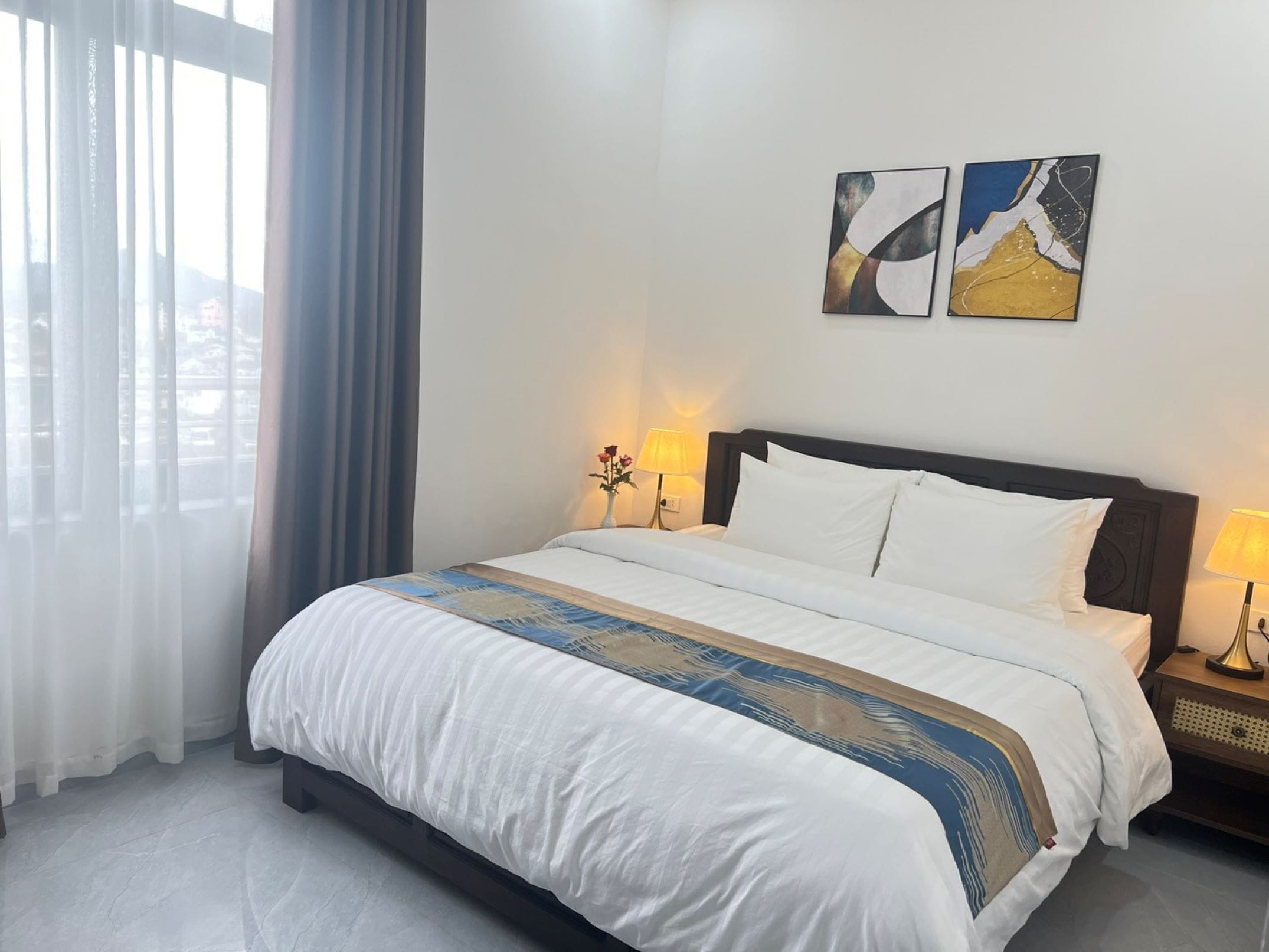 A hotel room with a large bed, two bedside lamps, a vase with flowers, and two abstract paintings on the wall.
