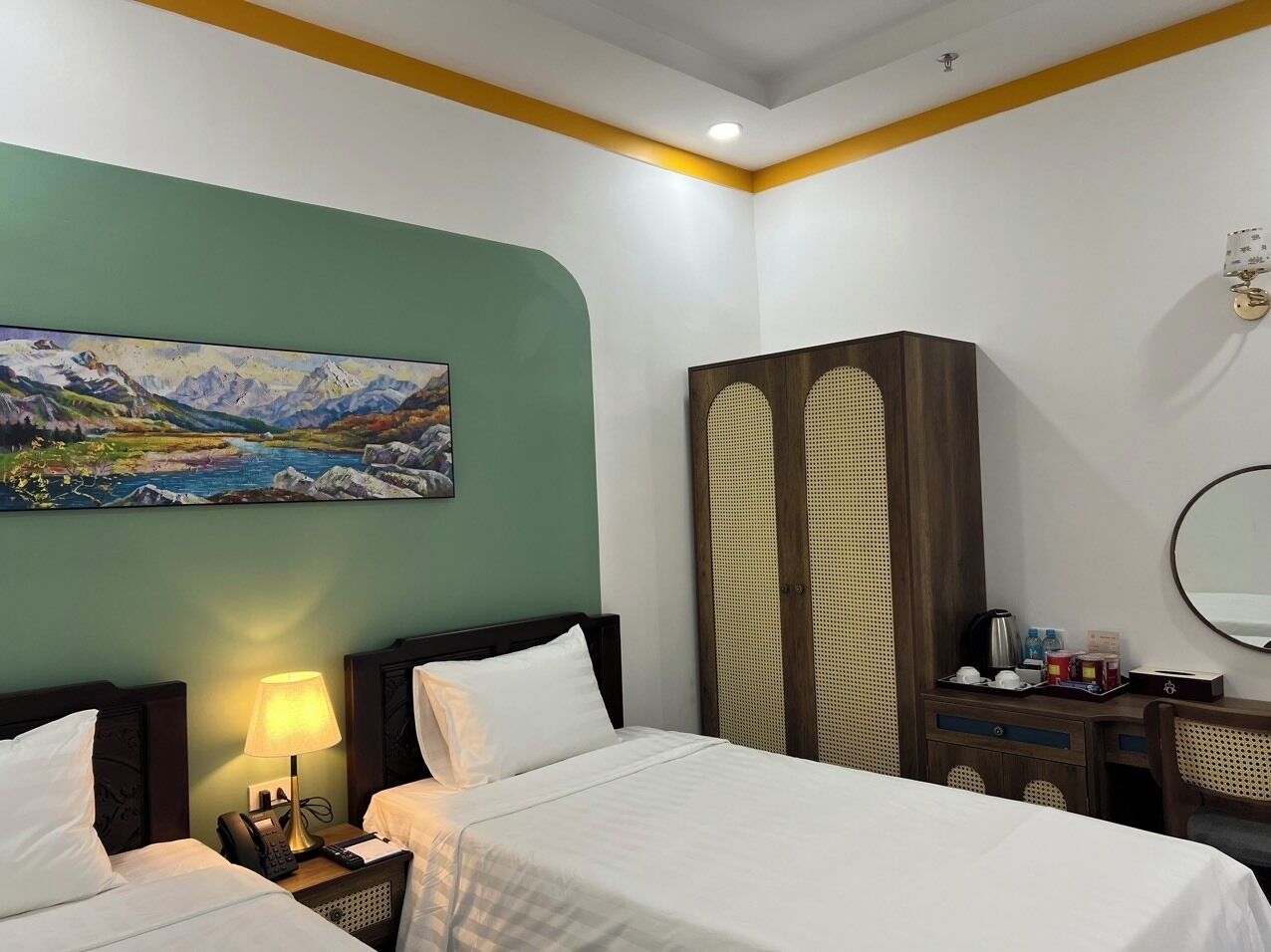 A hotel room with two beds, a painting of a mountain landscape, a wardrobe, a desk with a mirror, and a lamp.
