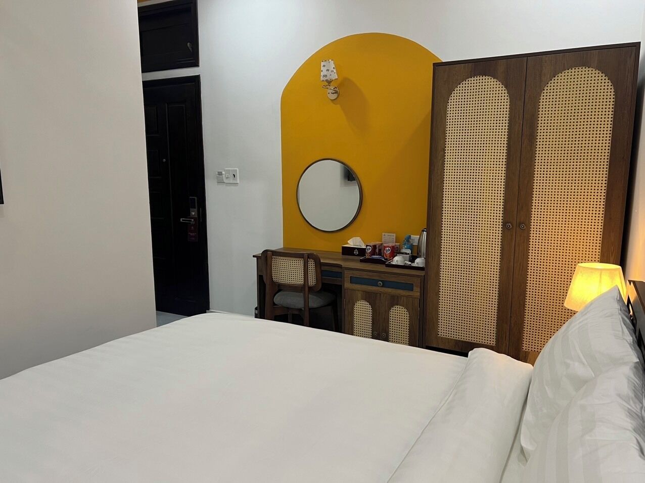 A hotel room with a bed, a desk with a chair, a round mirror, a yellow accent wall, and a wooden wardrobe with a woven pattern.