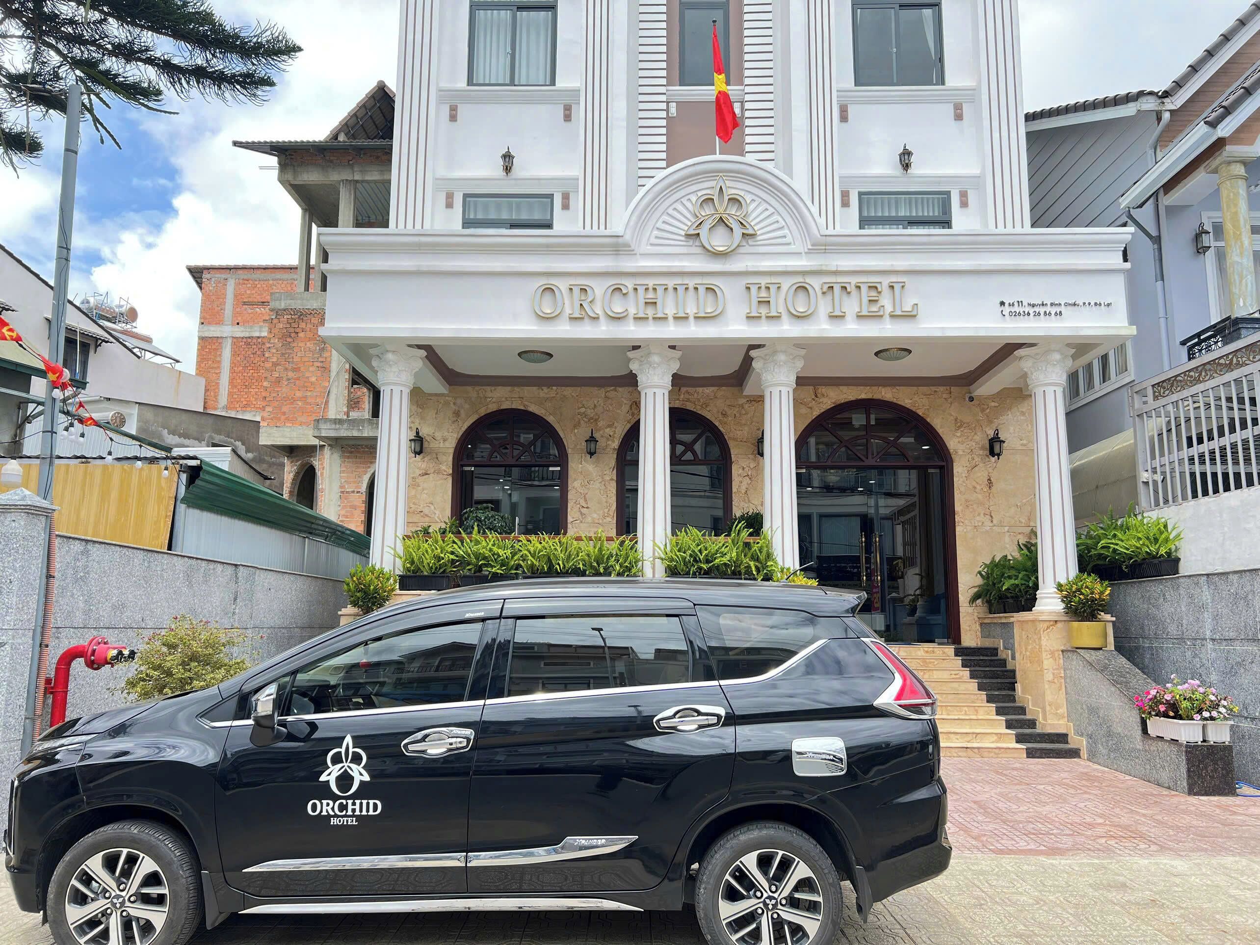 Exterior del Hotel Orchid, con una entrada destacada que incluye una bandera, una escalera que conduce a la entrada y un coche negro estacionado frente al hotel.