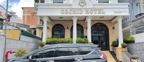 The exterior of the Orchid Hotel, featuring a prominent entrance with a flag, a staircase leading to the entrance, and a black car parked in front.