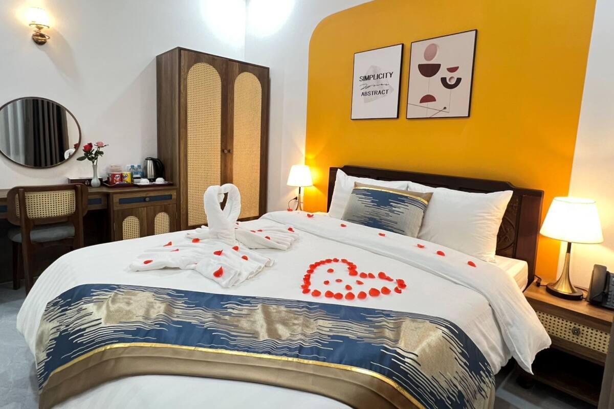 A hotel room with a bed, bedside lamps, a desk with a chair, a wardrobe, and two framed pictures on the wall.