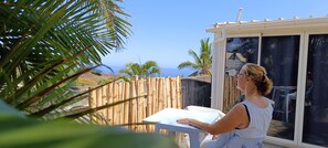 A person sitting at a table with a view of the ocean through a glass door. - Superb apartment, terrace, garden, 10 min. from beaches Saint-Gilles-Les-Bains (Saint-Paul)