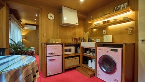 Een compacte keuken met een koelkast, magnetron en wasmachine, houten keukenkastjes en een klok aan de muur.