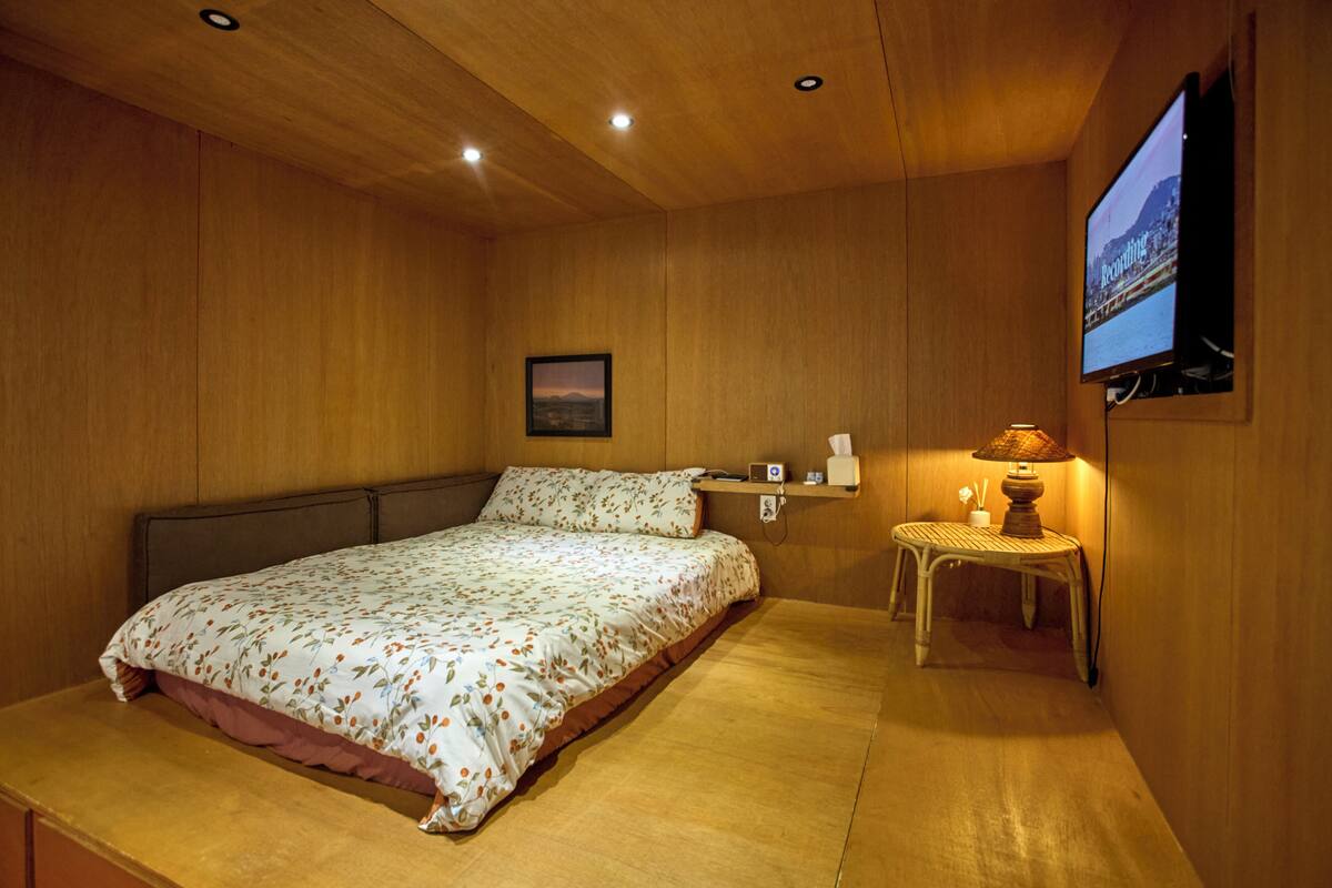 A bedroom with a bed, a bedside table with a lamp, a TV mounted on the wall, and a framed picture on the wall.