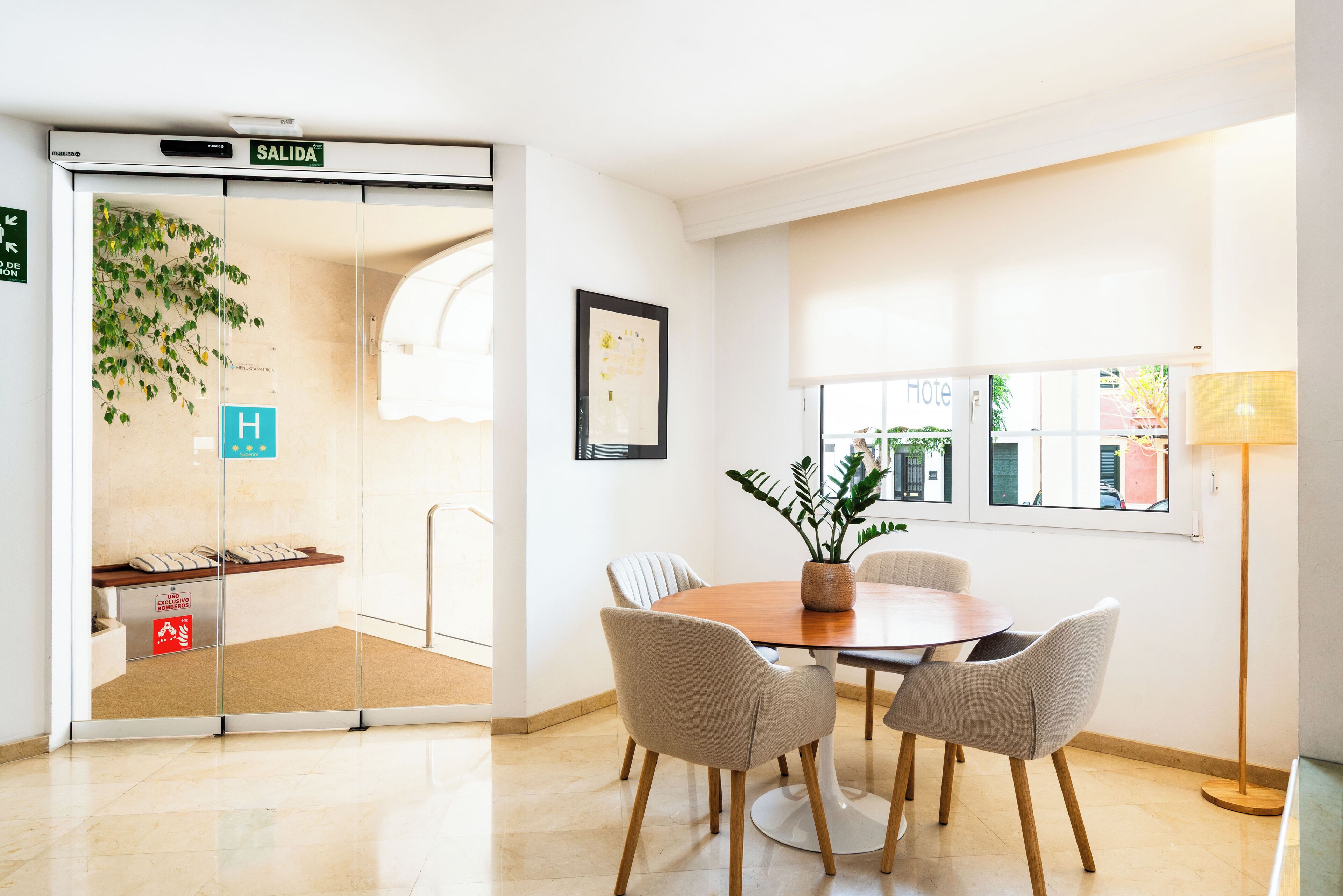 Habitación de hotel con una mesa de comedor de madera, cuatro sillas, una planta en maceta, un cuadro en la pared y una ventana con vistas al exterior.