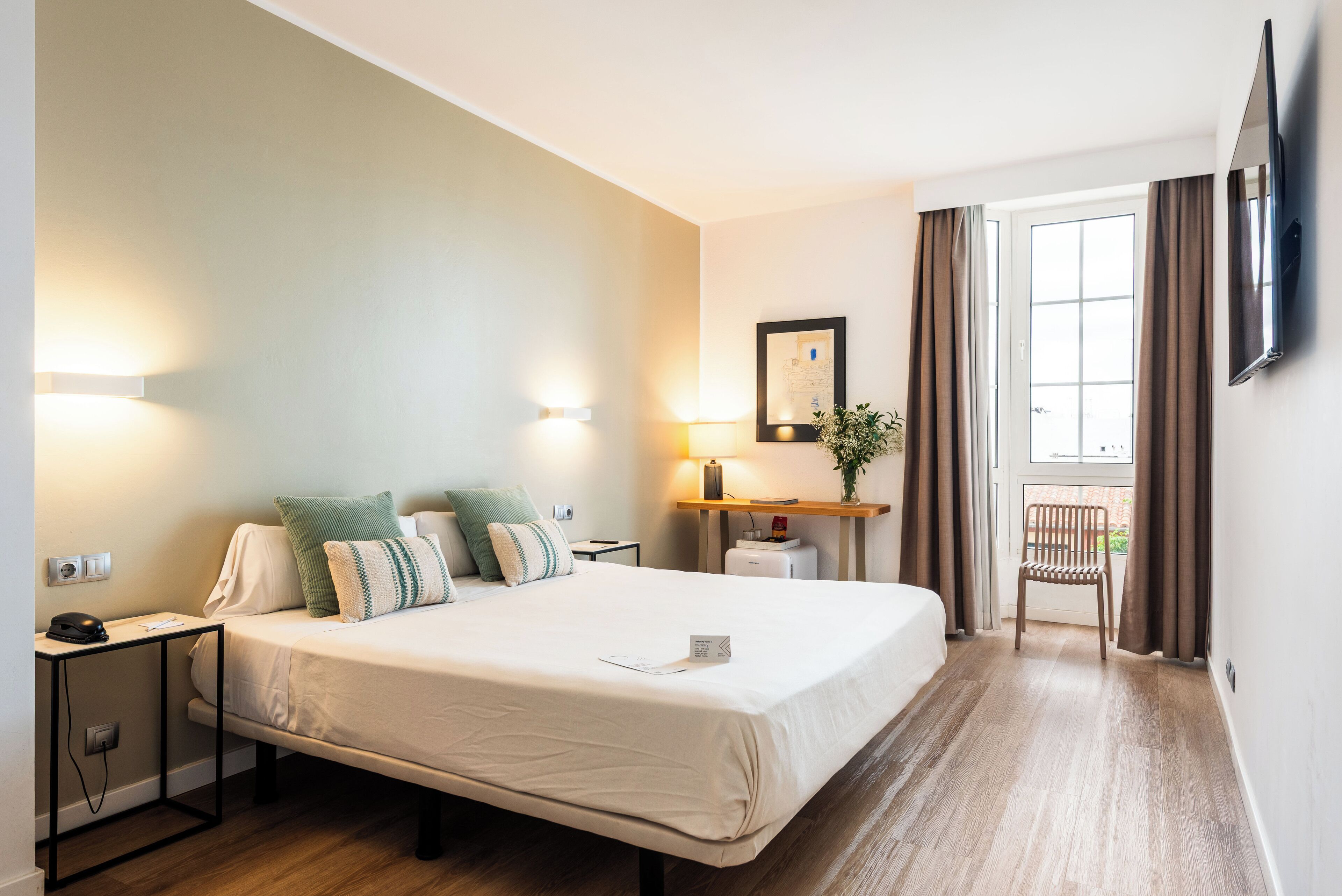 Habitación de hotel con cama, mesitas de noche, escritorio, silla, televisor y ventana con cortinas.