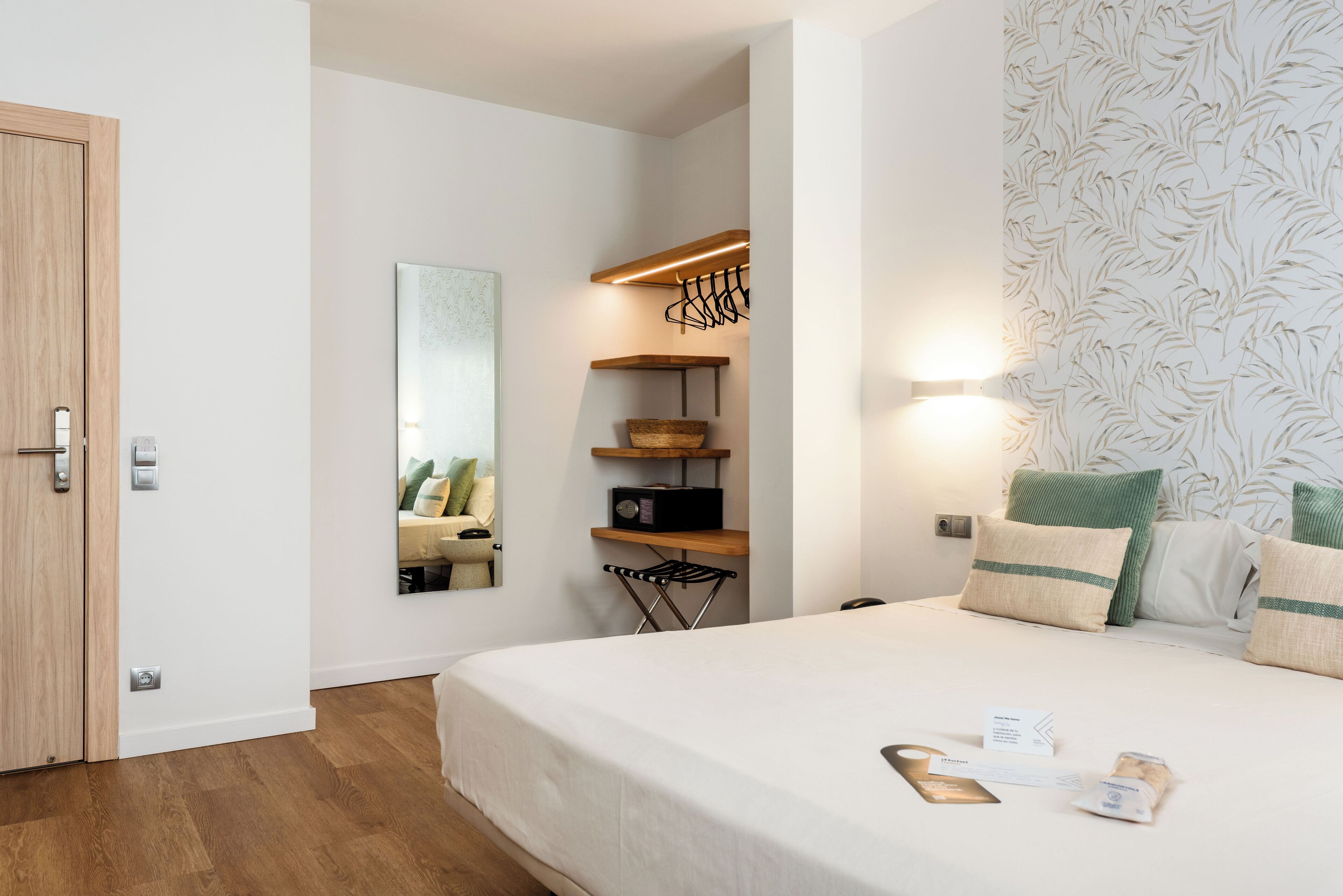 A hotel room with a wooden door, a bed with white bedding, a side table, a mirror, a shelf with a basket, and a patterned wallpaper.