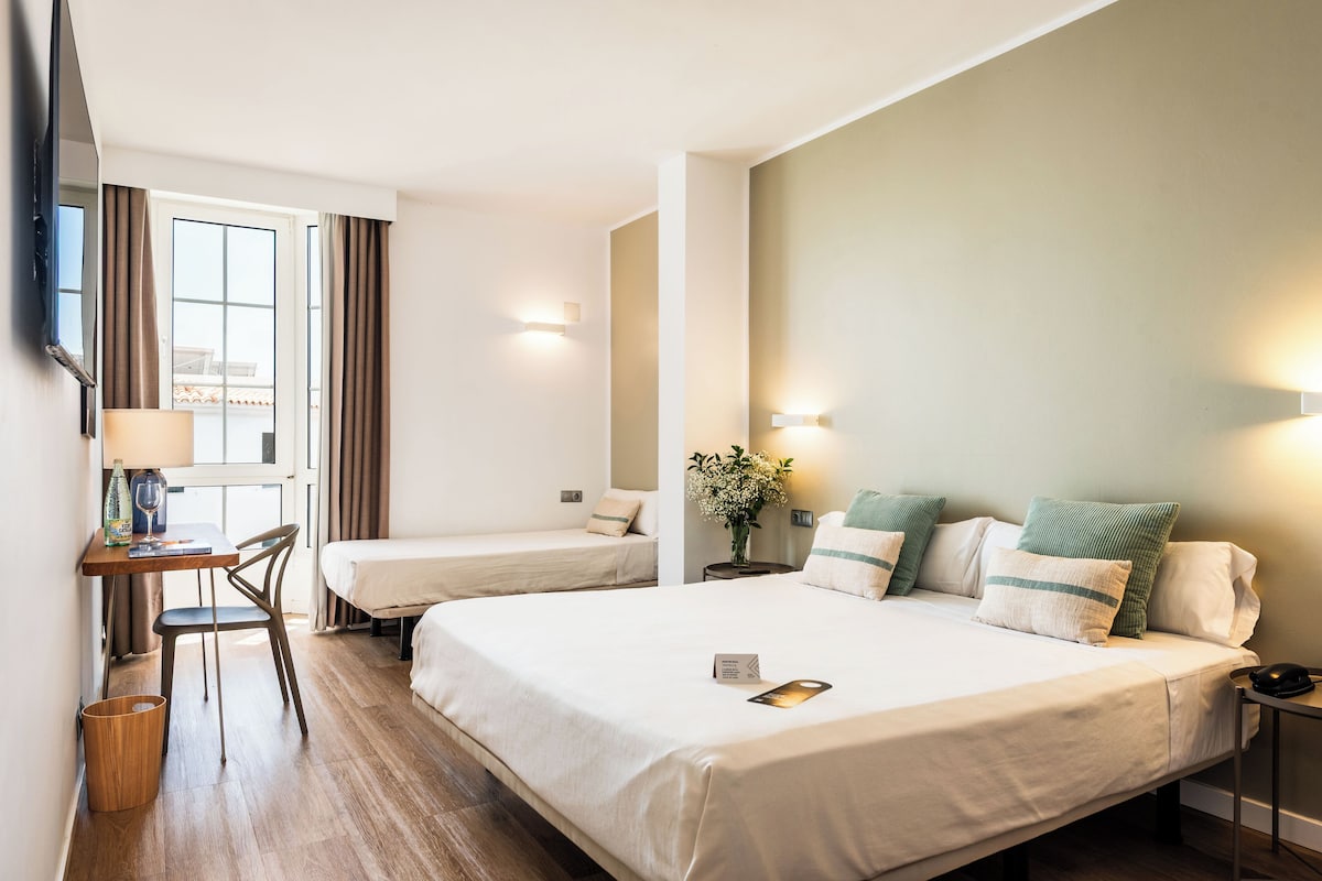 Ein Hotelzimmer mit zwei Betten, einem Schreibtisch mit Stuhl, einem kleinen Tisch mit Lampe und einem Fenster mit Vorhängen.