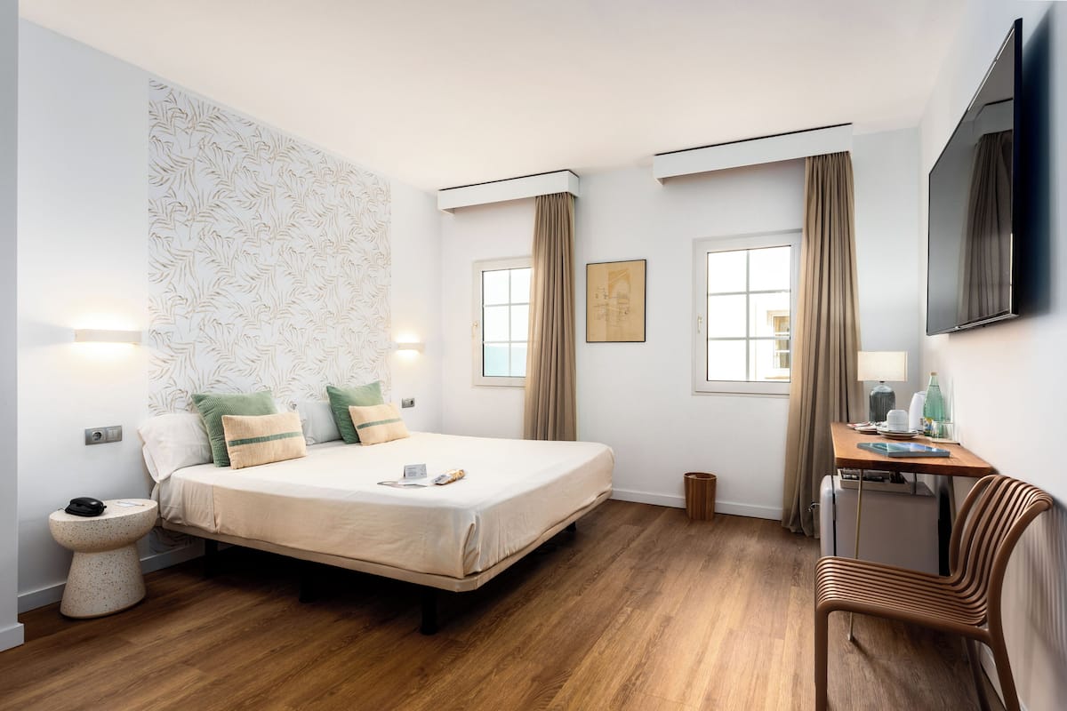 Ein Hotelzimmer mit Bett, Nachttisch, Lampe, Sessel und einem an der Wand befestigten Fernseher.