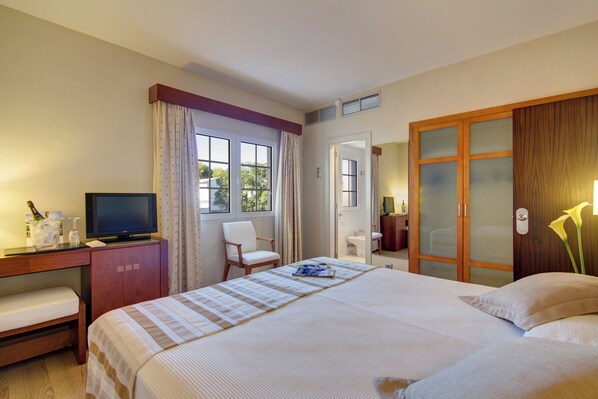 Habitación de hotel con cama, escritorio con televisor, silla, ventana con cortinas, baño y armario.