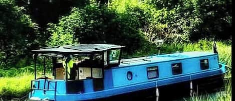 A narrowboat with a covered front and open sides, floating on water surrounded by greenery.
