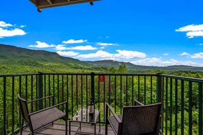 A balcony with a view of lush green mountains, a table with a bottle and glasses, and two chairs.