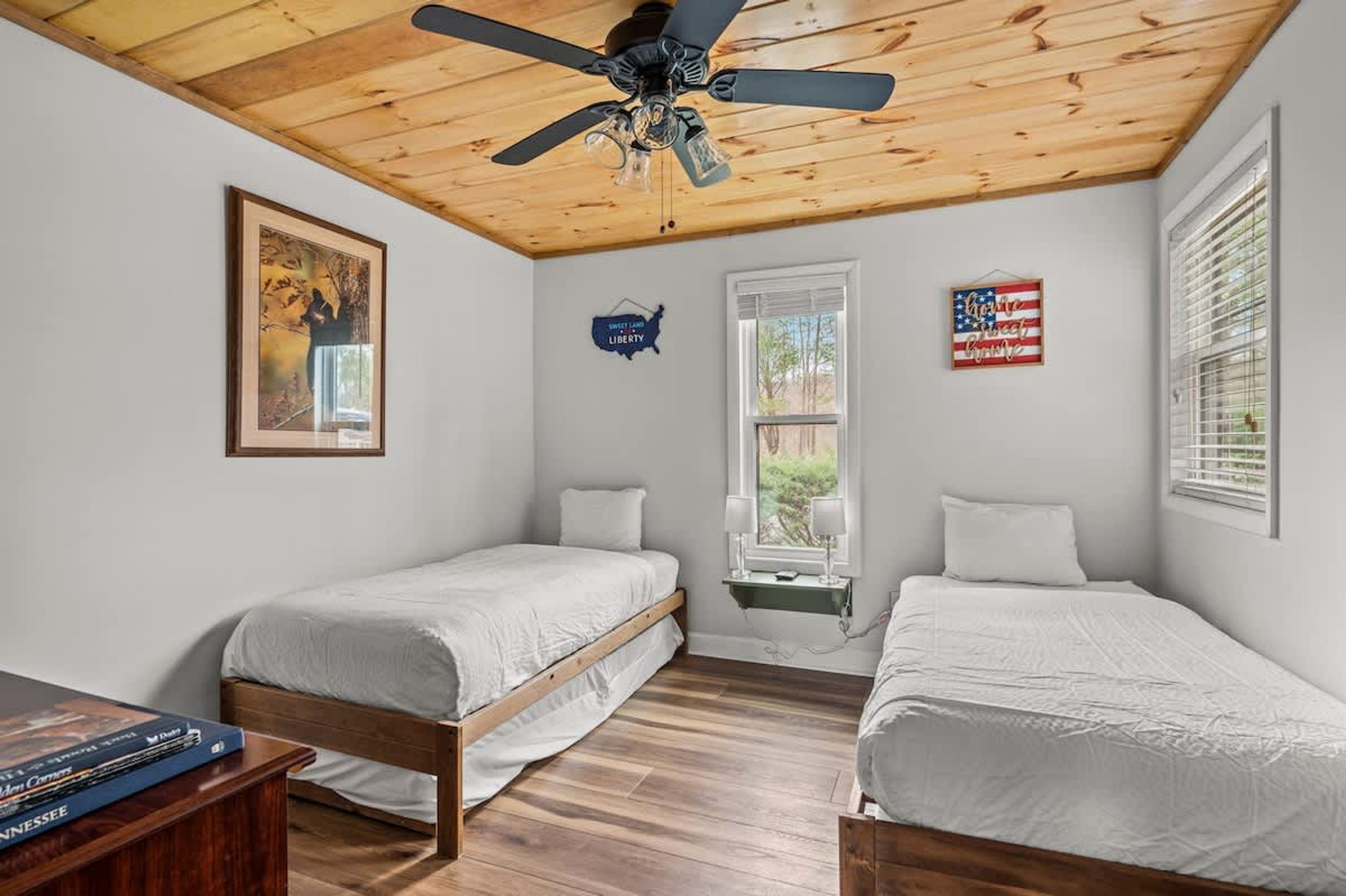 A bedroom with two single beds, wooden flooring, a ceiling fan, a window with blinds, and wall decorations including a map and an American flag sign.
