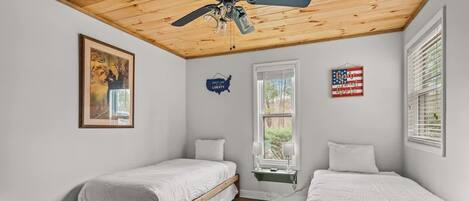 A bedroom with two single beds, wooden flooring, a ceiling fan, a window with blinds, and wall decorations including a map and an American flag sign.