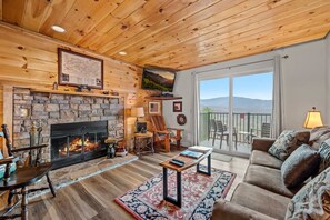 A cozy living room with a stone fireplace, wooden ceiling, and a balcony with outdoor seating. - Indoor Pool Sleeps 7 Mountain Views Playground (Gatlinburg)