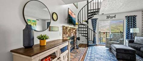 A modern living room with a wooden cabinet, a round mirror, a staircase leading to a balcony, and a view of the outdoors.