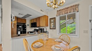 Un coin repas avec une table en bois dressée pour quatre personnes, une cuisine en arrière-plan et une fenêtre avec des stores.