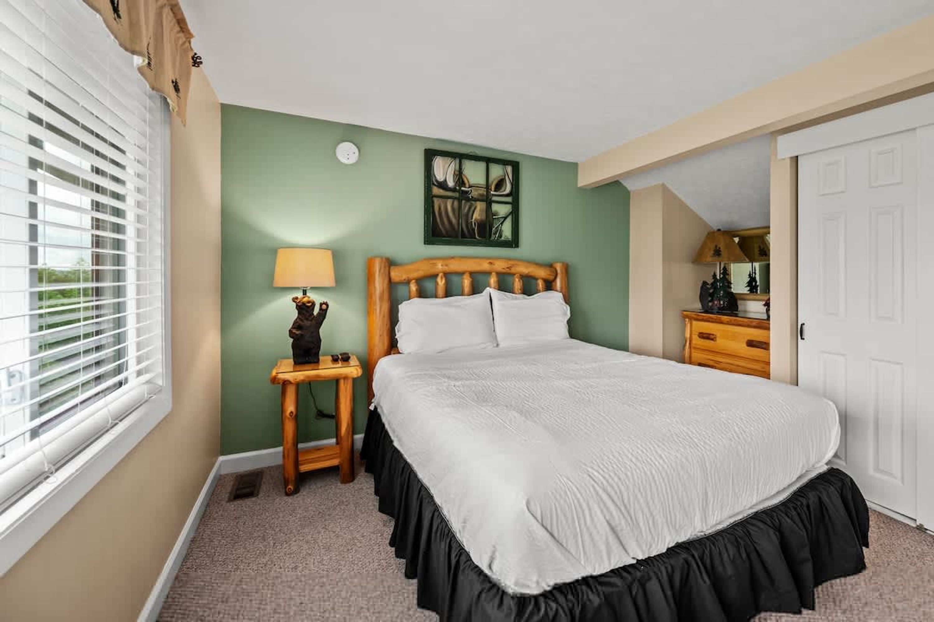 A bedroom with a bed, a nightstand, a lamp, a mirror, and a window with blinds.