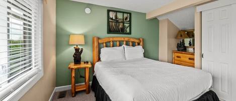 A bedroom with a bed, a nightstand, a lamp, a mirror, and a window with blinds.