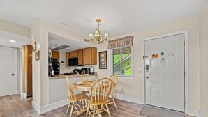 Un coin repas avec une table et des chaises en bois, une cuisine avec des placards en bois et une porte blanche avec un paillasson.