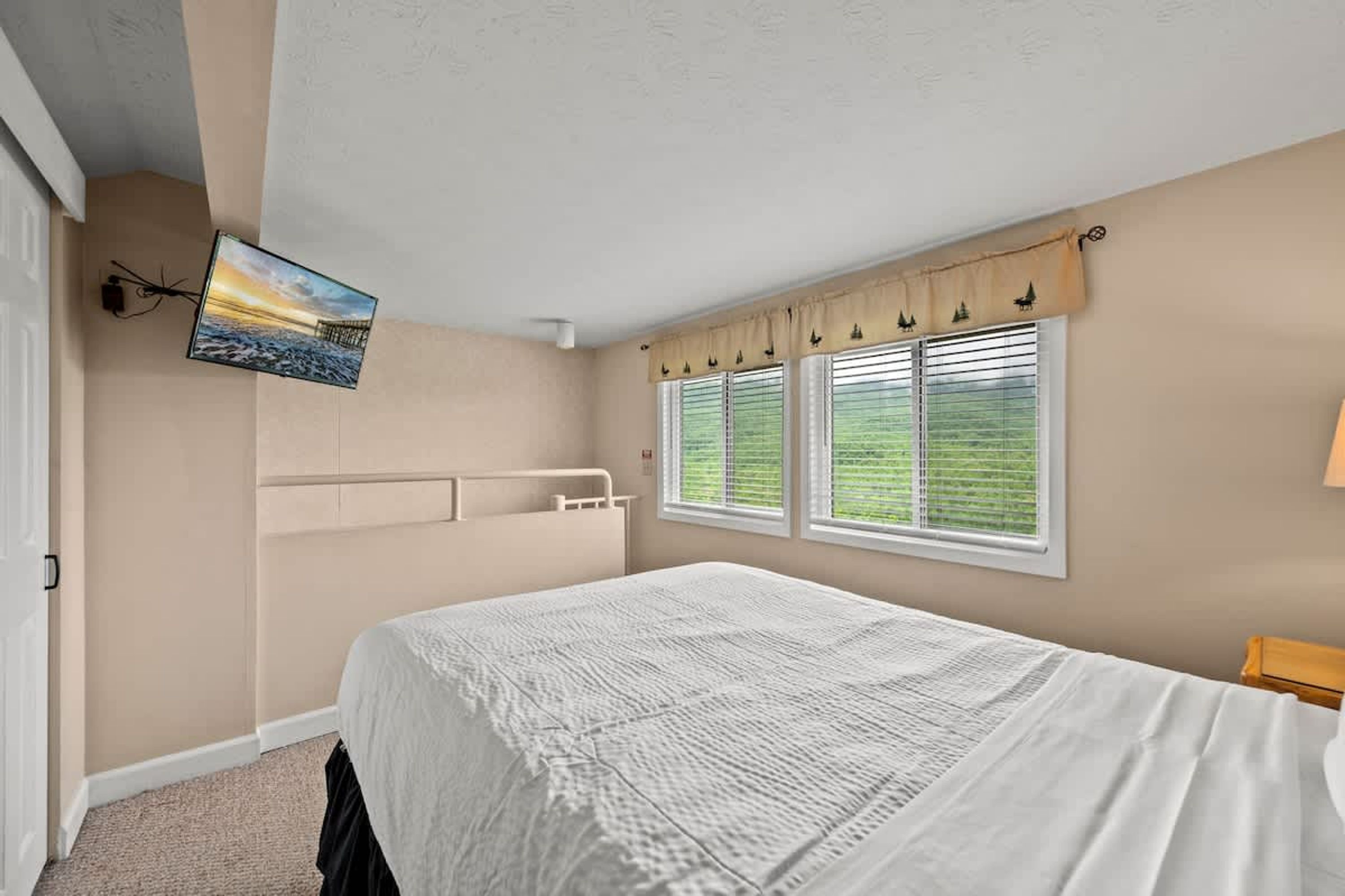 Ein Schlafzimmer mit einem Bett, einem an der Wand befestigten Fernseher und einem Fenster mit Jalousien.