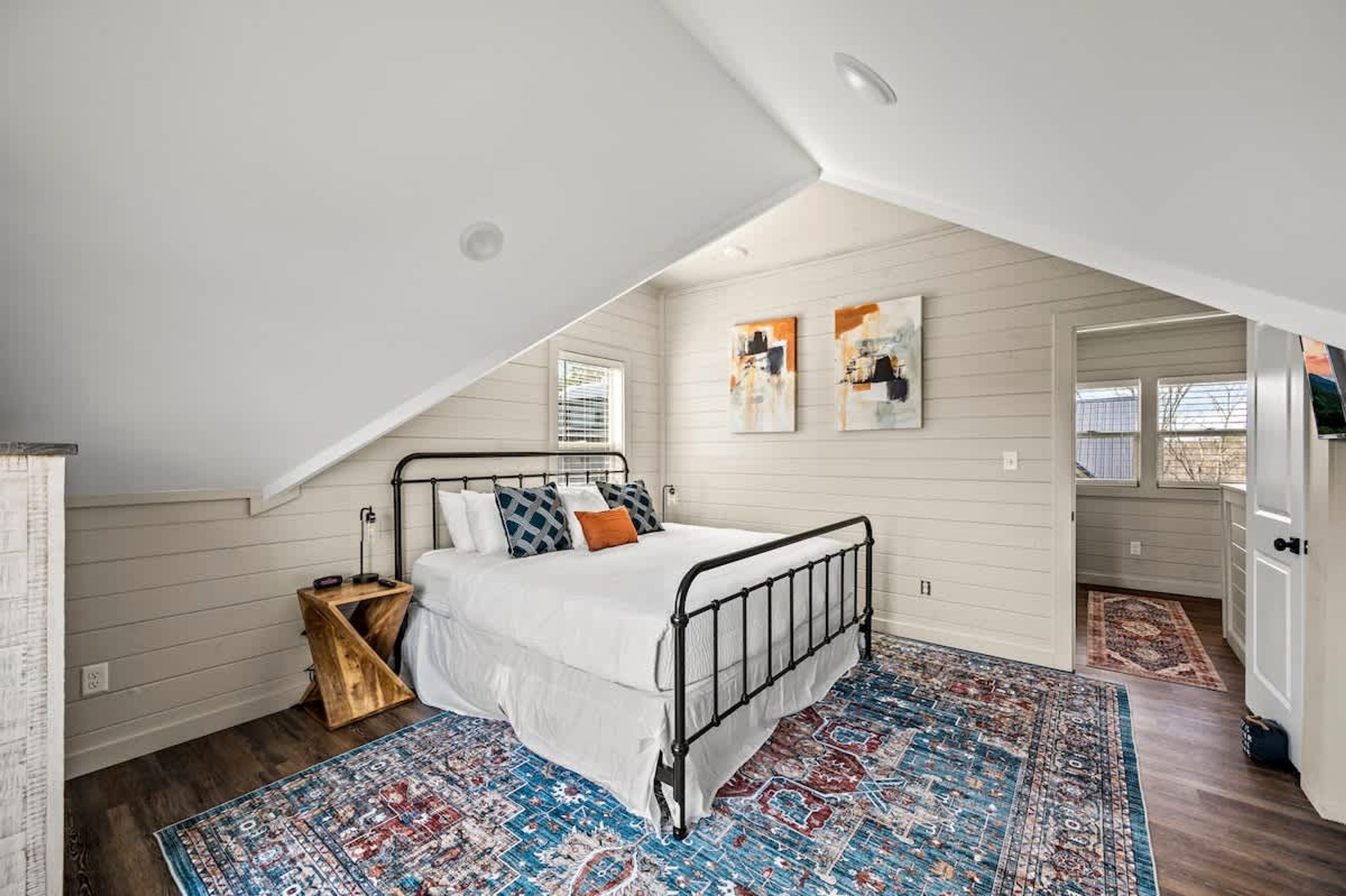 A bedroom with a sloped ceiling, a bed with a white bedspread and multiple pillows, a patterned rug, and a small wooden side table.