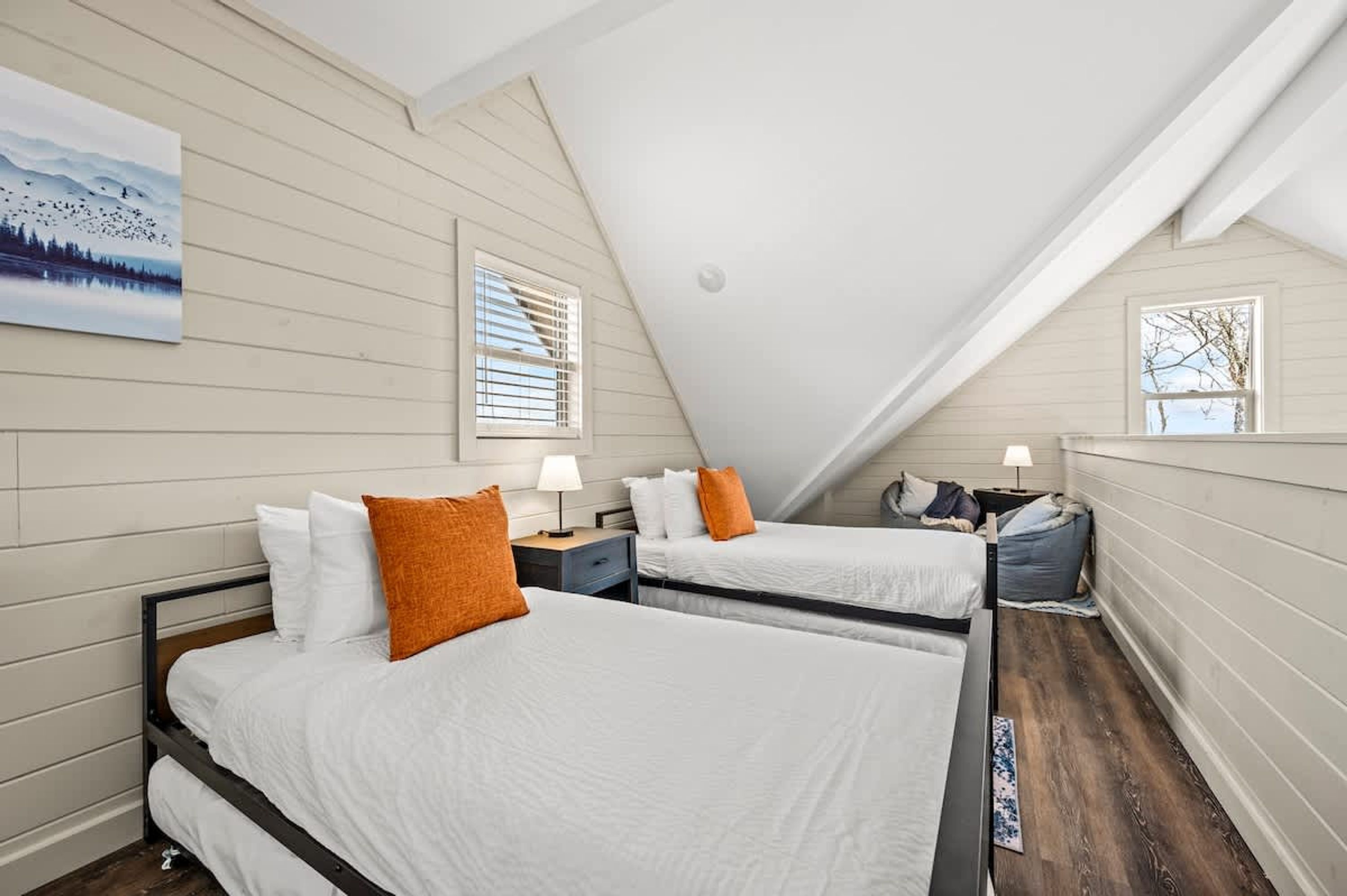 A bedroom with two beds, white bedding, and orange pillows. There is a painting on the wall, a window with blinds, and a lamp on the nightstand.
