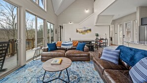 A spacious living area with a brown leather sofa, a round coffee table, and a blue patterned rug. There is a dining table with chairs, a kitchen area with a refrigerator, and large windows offering a view of the outdoors. - NEW Lake View Indoor Pool Sleeps 14 Hot Tub (Sevierville)