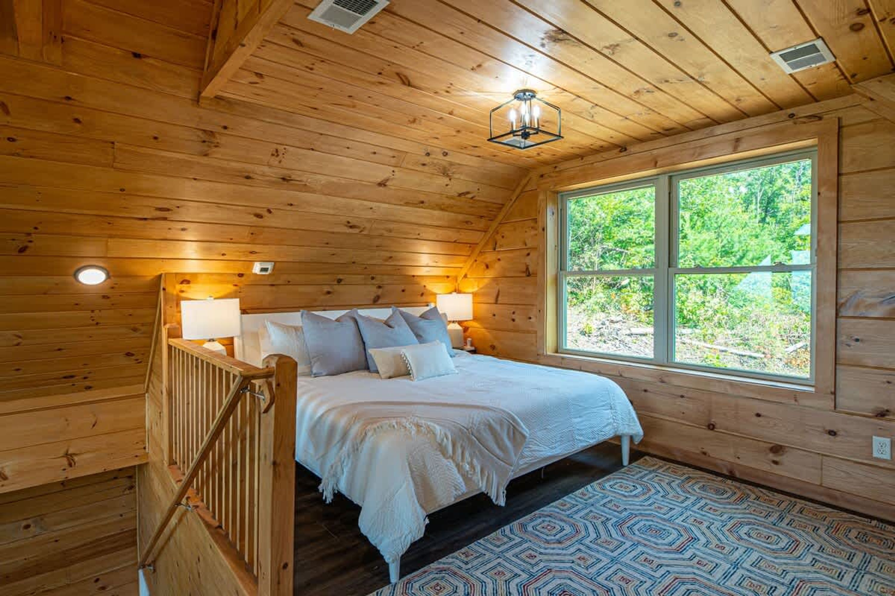 A wooden cabin bedroom with a bed, two bedside lamps, a window with a view of greenery, and a patterned rug.