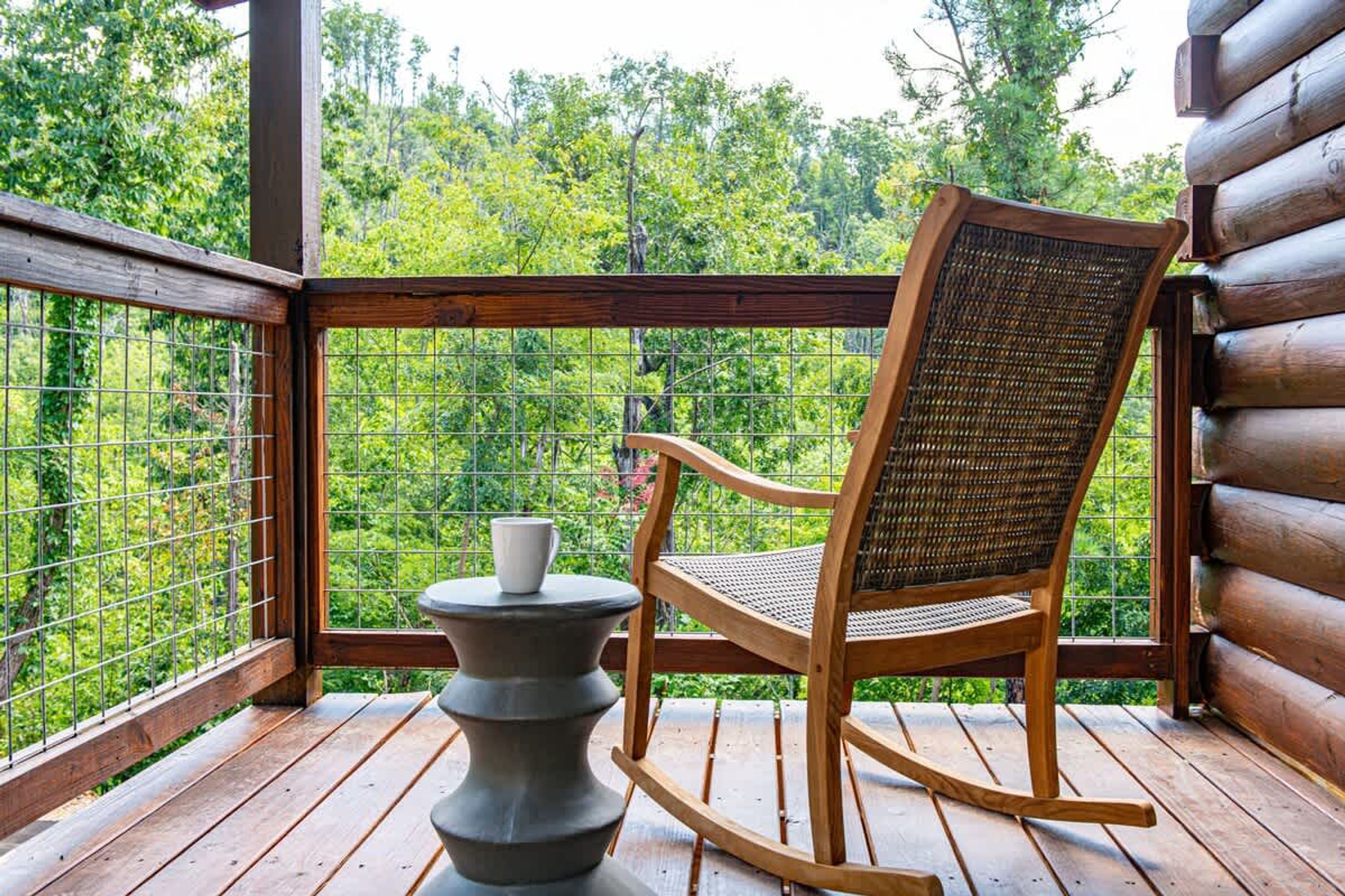 A wooden rocking chair on a wooden deck with a metal railing, overlooking a forested area.
