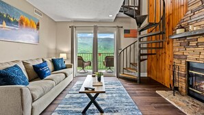 A living room with a sofa, coffee table, and a fireplace. There is a staircase leading to a balcony with a view of the mountains.
