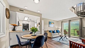 A modern kitchen with white cabinets, a dining area with a round table and chairs, a living room with a sofa and a view of the outdoors.