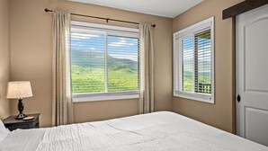 A bedroom with a large bed, two windows with curtains, a nightstand with a lamp, and a view of green hills.