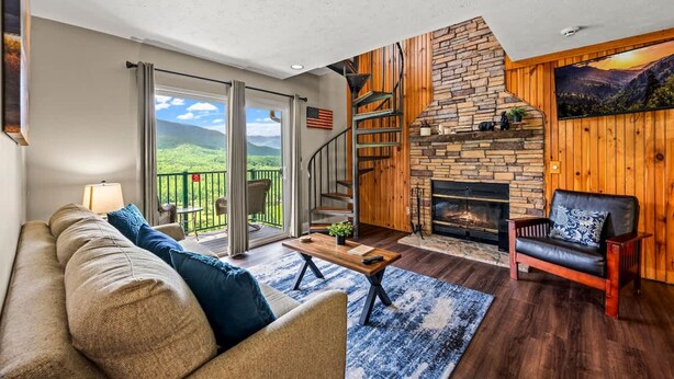 A living room with a stone fireplace, a wooden staircase, and a balcony with a view of the mountains.