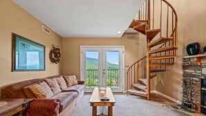 A living room with a brown leather sofa, a wooden coffee table, a spiral staircase, and a stone fireplace. - Hot Tub Heated Pool Mountain Views Sleeps 6 (Gatlinburg)