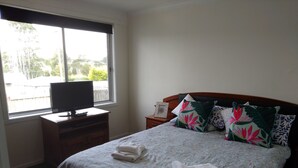 Un dormitorio con cama, televisor sobre un mueble de madera, ventana con vista a follaje y mesita de noche con lámpara.