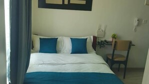Un lit bien fait, recouvert d’une couverture bleue et blanche, une œuvre d’art accrochée au mur, un bureau avec une chaise et une petite plante en pot.