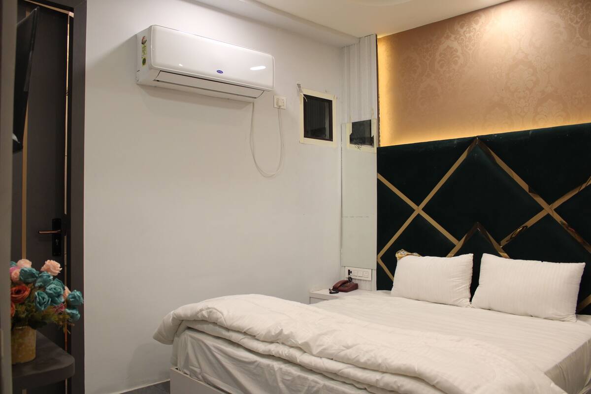 A hotel room with a bed, a bedside table, a telephone, a wall-mounted air conditioner, a small window, and a decorative wall panel.