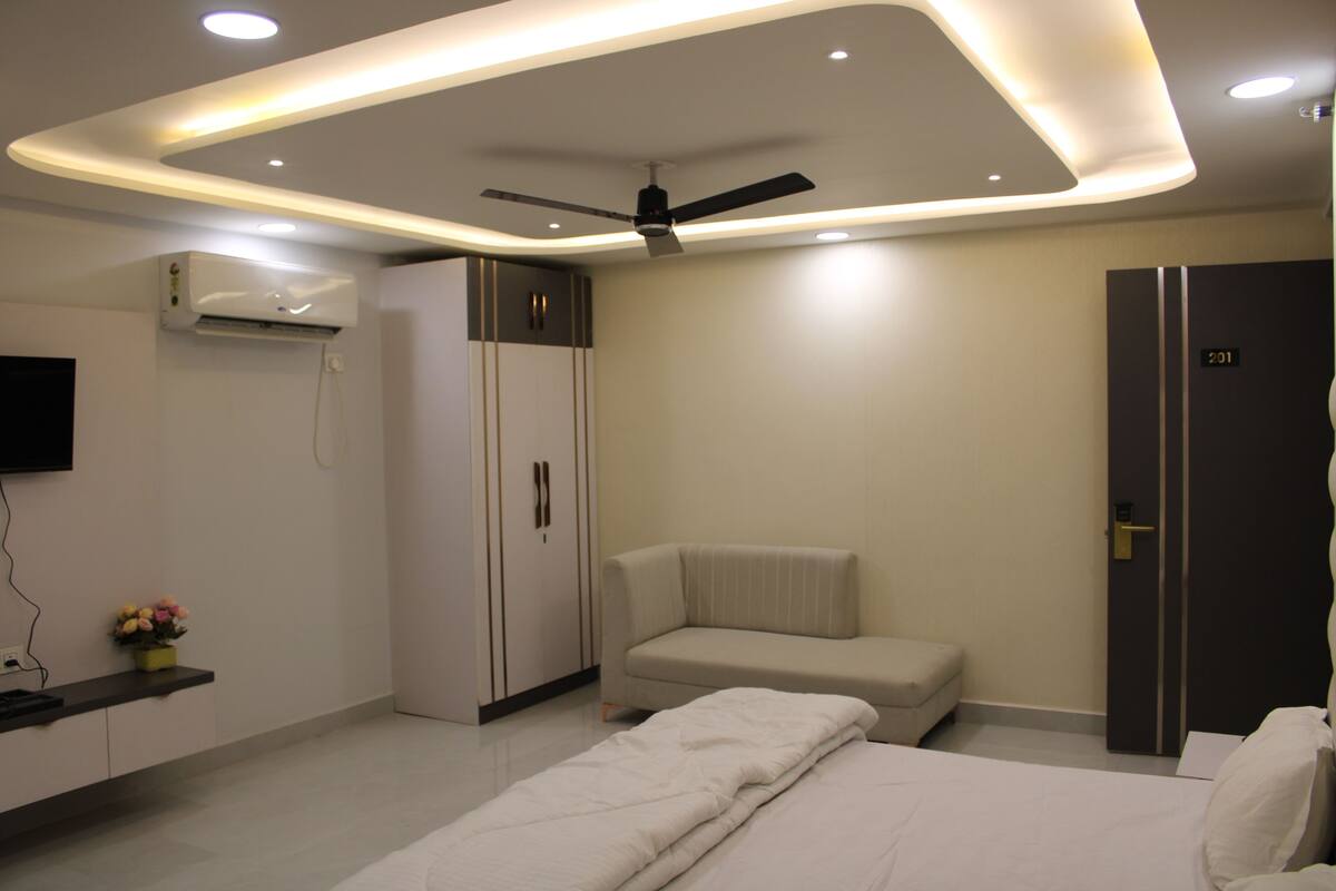 A modern hotel room with a bed, a sofa, a ceiling fan, recessed lighting, and an air conditioning unit.