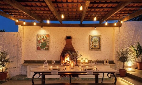 Un espace extérieur aménagé pour les repas, avec une cheminée en briques, des plantes en pot et des guirlandes lumineuses.
