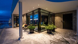 Un edificio moderno con techo curvo, amplios ventanales de vidrio y pared de piedra. Se observan plantas en macetas y mobiliario exterior.