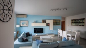 A modern living room with a blue wall, a large clock, a flat-screen TV, a dining area with white chairs, and a coffee table.
