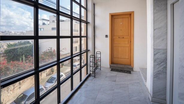 A corridor with large windows, a wooden door, and a tiled floor.