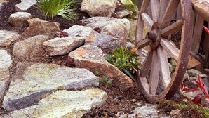 A rustic garden with a wooden fence, a wooden cart wheel, and various rocks and plants.